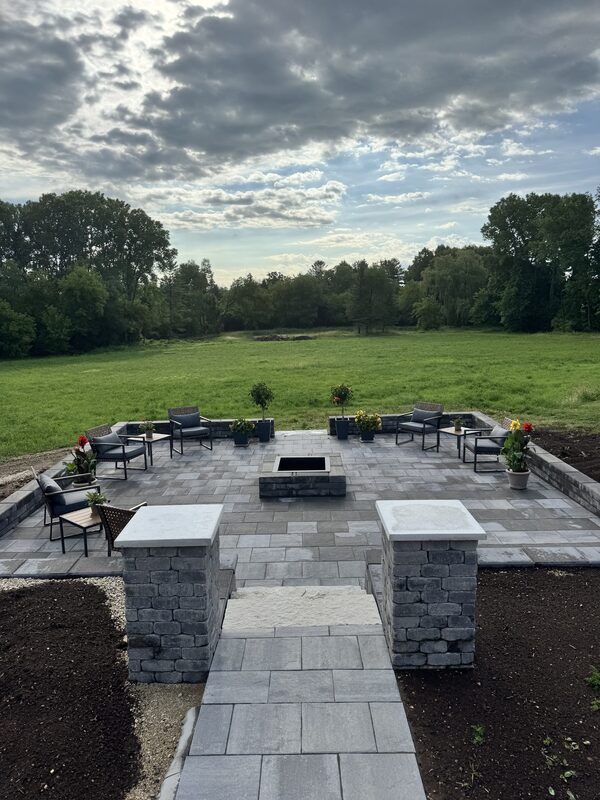 Large paver patio with a central fire pit, stone pillars and modern outdoor furniture overlooking open fields